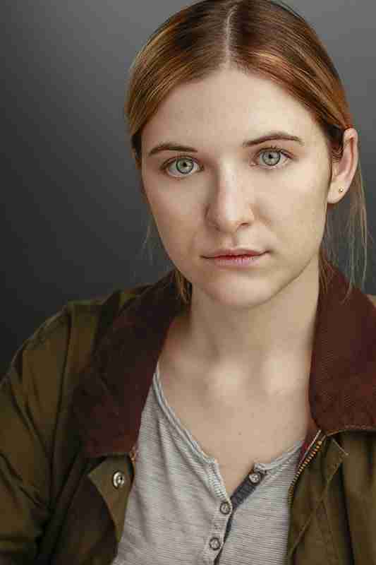Actor headshot: Alex Hannant, golden-brown hair pulled back, green eyes and freckles, olive-green jacket over a striped henley, grey studio backdrop.