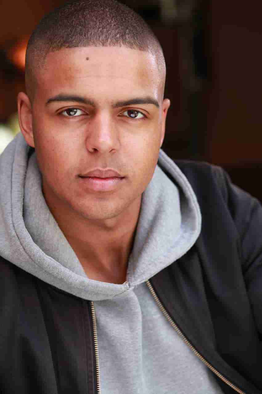 Actor headshot: Kofi Dennis, short buzz cut, serious expression, grey hoodie under a black bomber jacket, dark blurred background.