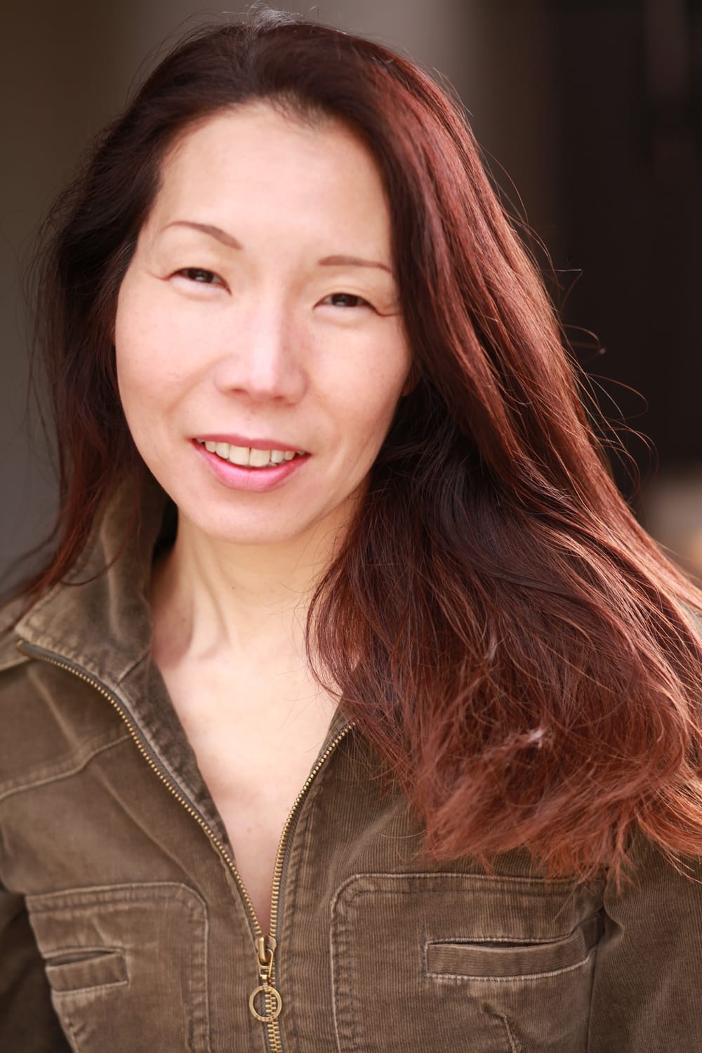 Actor headshot: Meg Kabota, long auburn hair swept over shoulder, warm smile, olive-brown corduroy jacket.