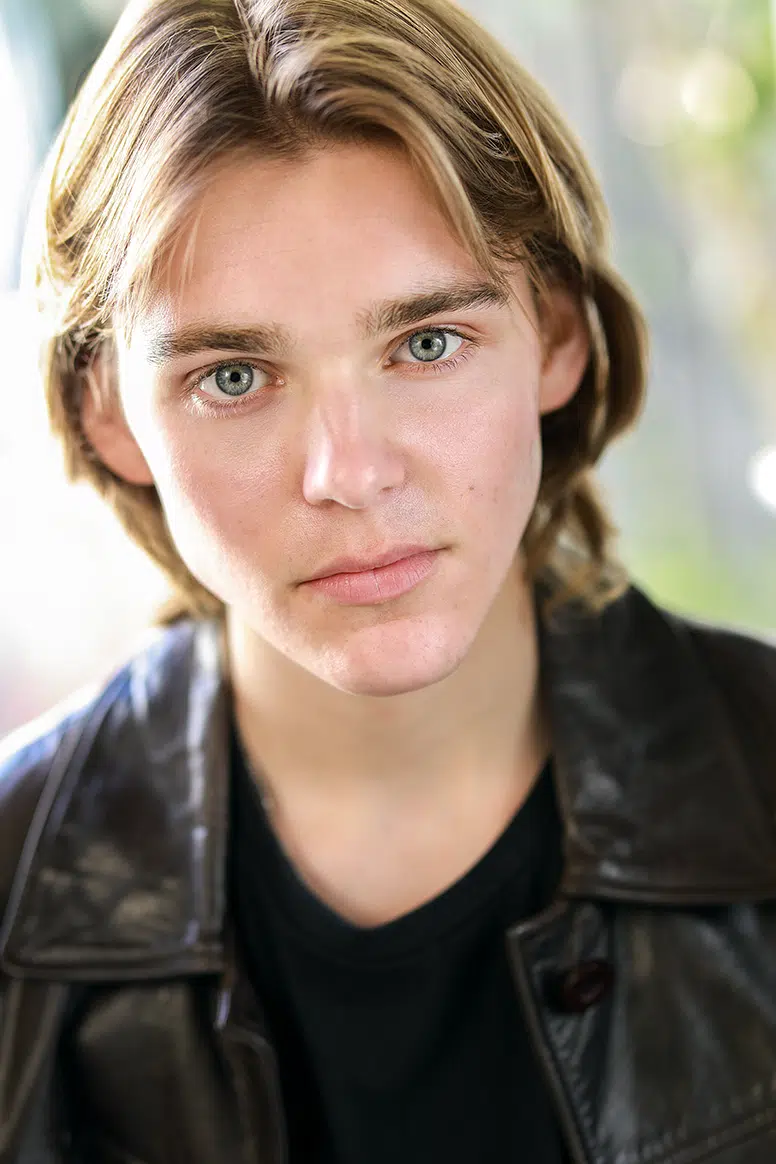Actor headshot: Will Wickham, shoulder-length wavy light brown hair, pale blue-grey eyes, silver chain necklace, brown leather jacket over a light blue v-neck sweater, charcoal grey studio backdrop.
